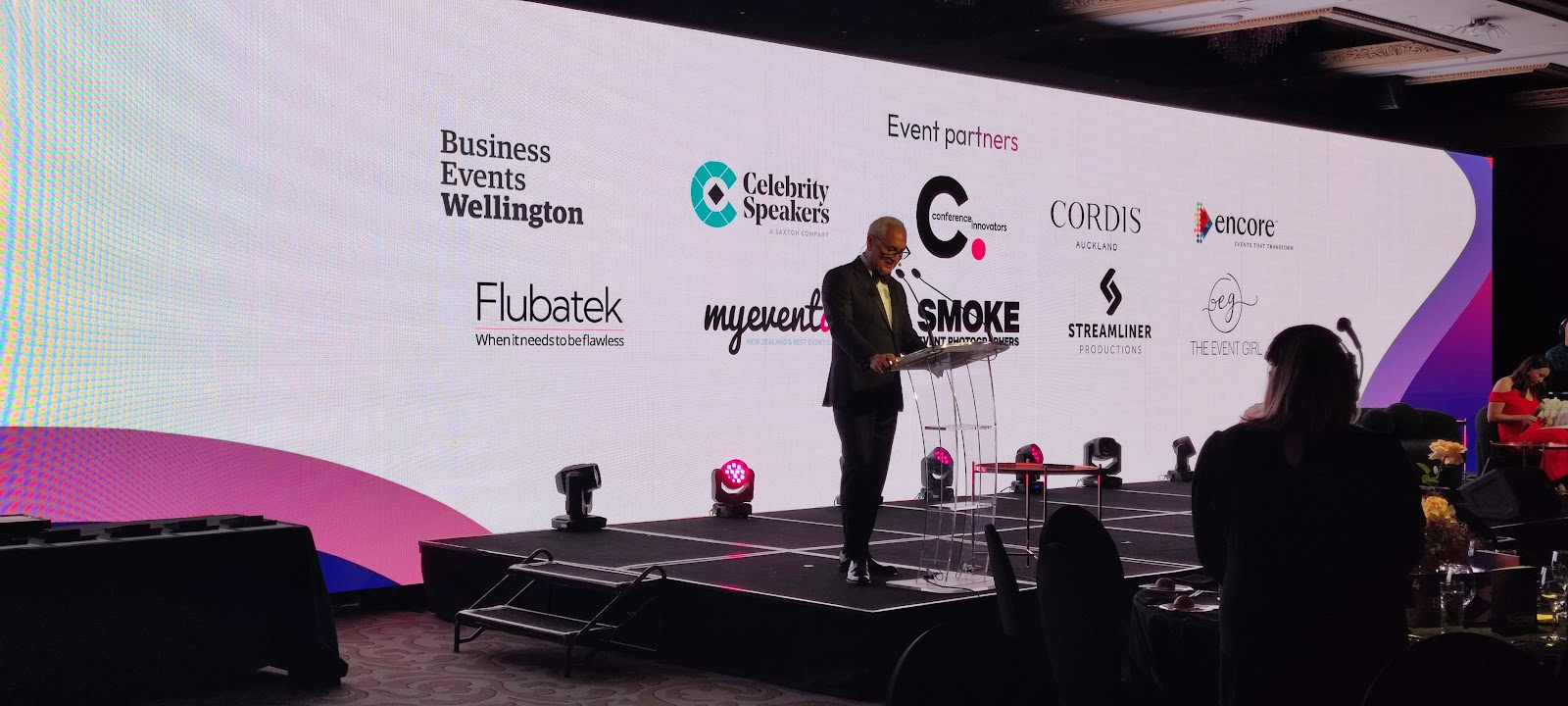 Speaker presenting on stage in front of a large event partner screen produced by Flubatek.